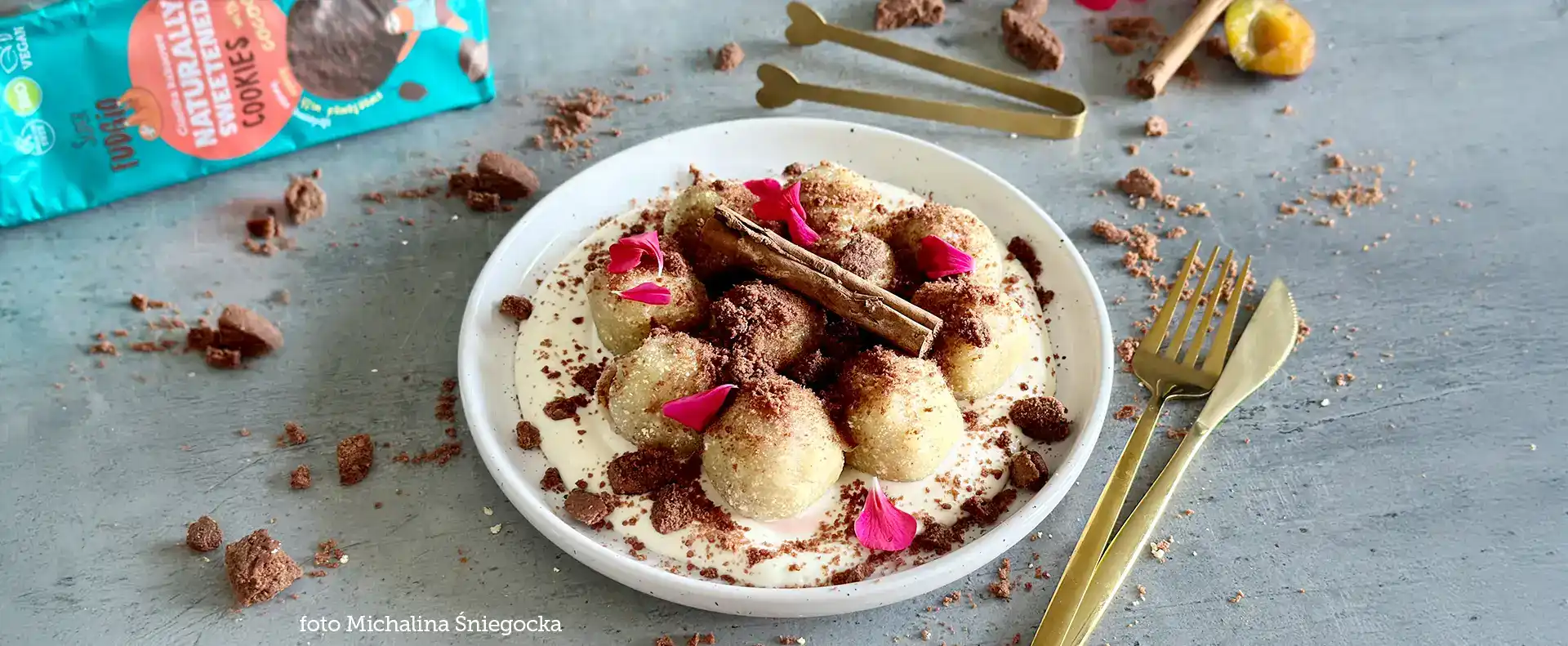 wegańskie knedle z tofu ze śliwkami i ciasteczkową kruszonką, podane na białym talerzu, udekorowane cynamonem i płatkami róży, wyeksponowane na szarym blacie w tle niebieskie opakowanie ciastek