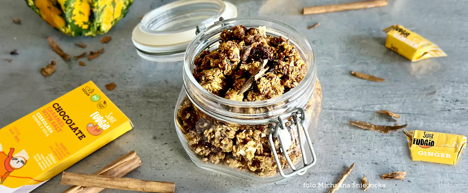 granola dyniowa w słoiku ze szklaną pokrywką na szarym blacie; wokół krówki imbirowe, cynamon, czekolada w żółtym opakowaniu